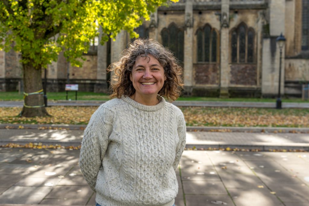 A photo of Green Councillor Lisa Stone, standing outside a building under a tree. She is smiling and wearing a white jumper.